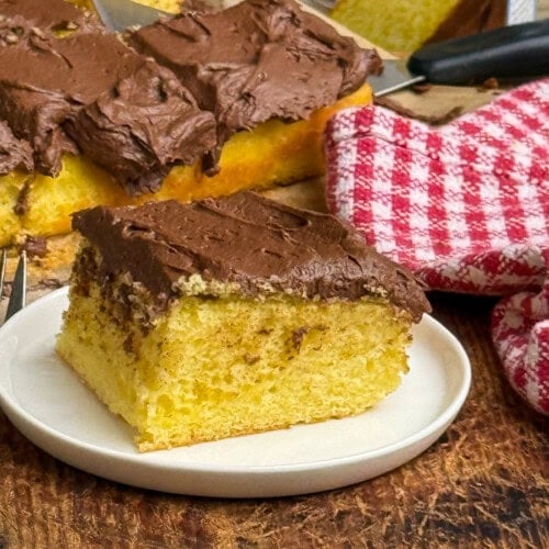 A slice of yellow sheet cake with chocolate frosting sits on a white plate, with the remaining cake and a red-and-white checkered cloth visible in the background.