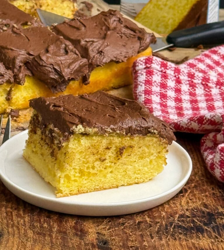 A slice of yellow sheet cake with chocolate frosting sits on a white plate, with the remaining cake and a red-and-white checkered cloth visible in the background.