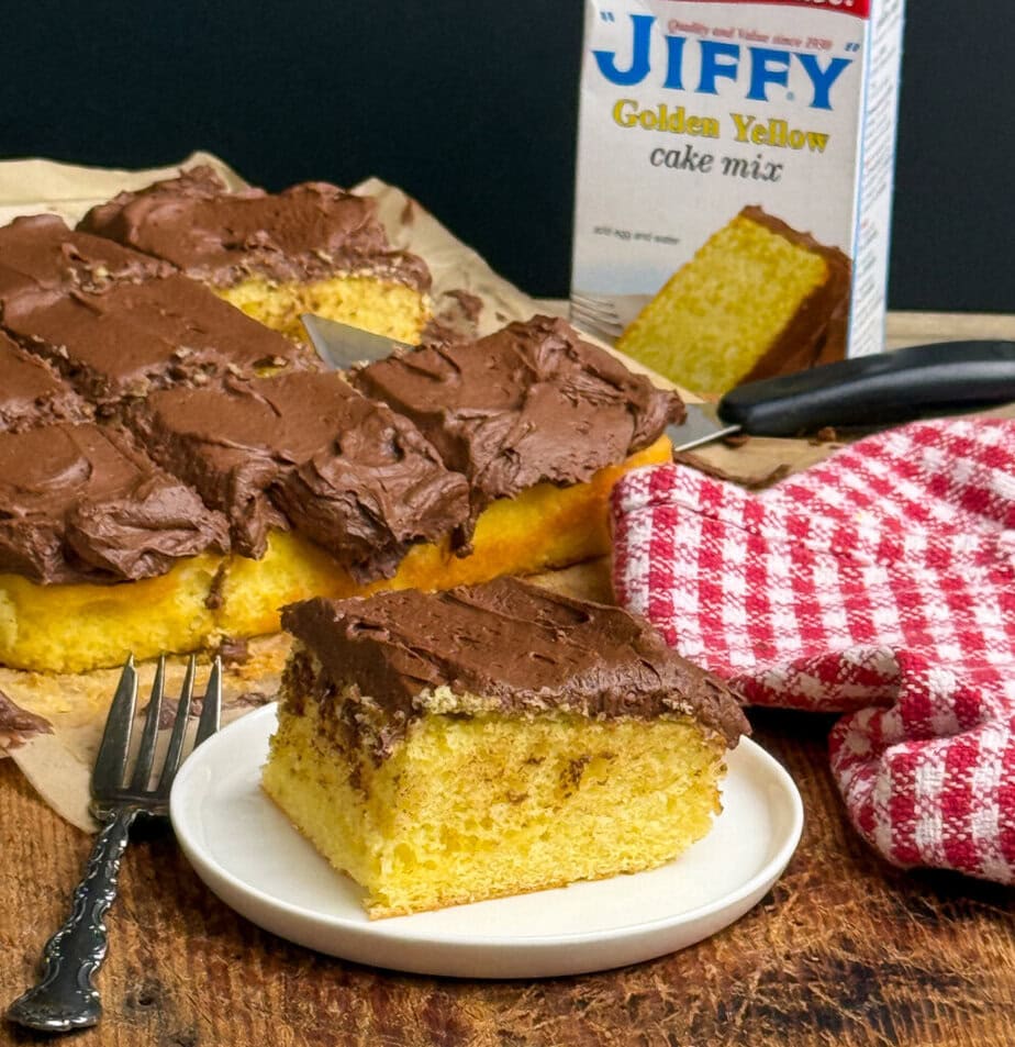 A square slice of yellow cake with chocolate frosting sits on a plate in front of the rest of the cake. The cake is on parchment paper, next to a red checkered towel and a Jiffy Golden Yellow cake mix box.