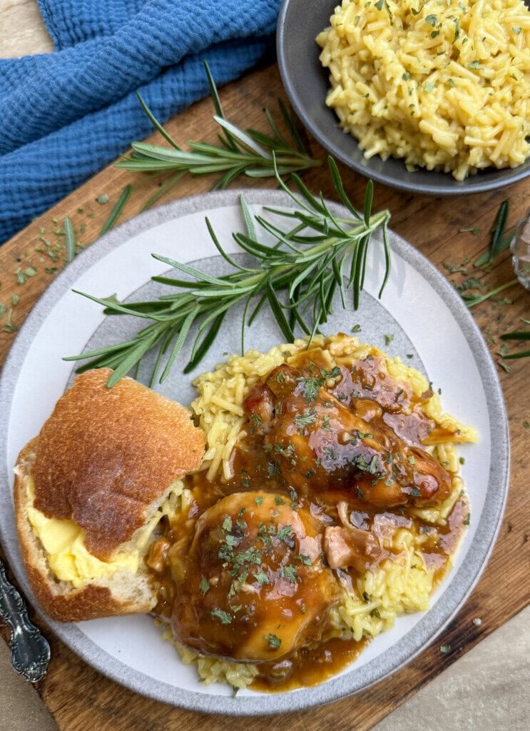 A plate with two pieces of smothered chicken in brown gravy over yellow rice, garnished with herbs, served with a buttered roll. A sprig of rosemary and additional rice in a bowl are nearby on a wooden surface.