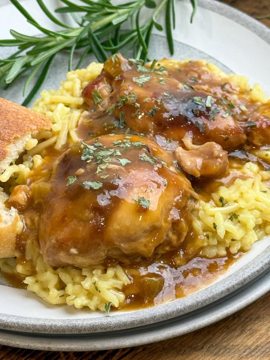 A plate of yellow rice topped with saucy chicken thighs garnished with herbs, served with a slice of bread and a sprig of fresh rosemary on the side.