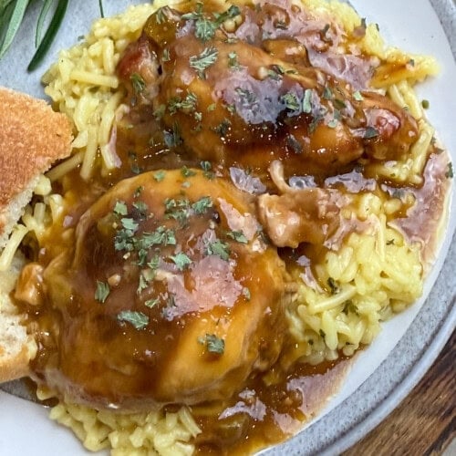 A plate of yellow rice topped with two pieces of smothered chicken in brown gravy, garnished with herbs. A slice of bread and sprigs of fresh rosemary are on the side.