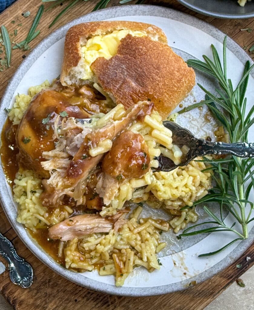 A plate with shredded roasted chicken in gravy over yellow rice, served with a buttered bread roll and garnished with fresh rosemary on a wooden table.