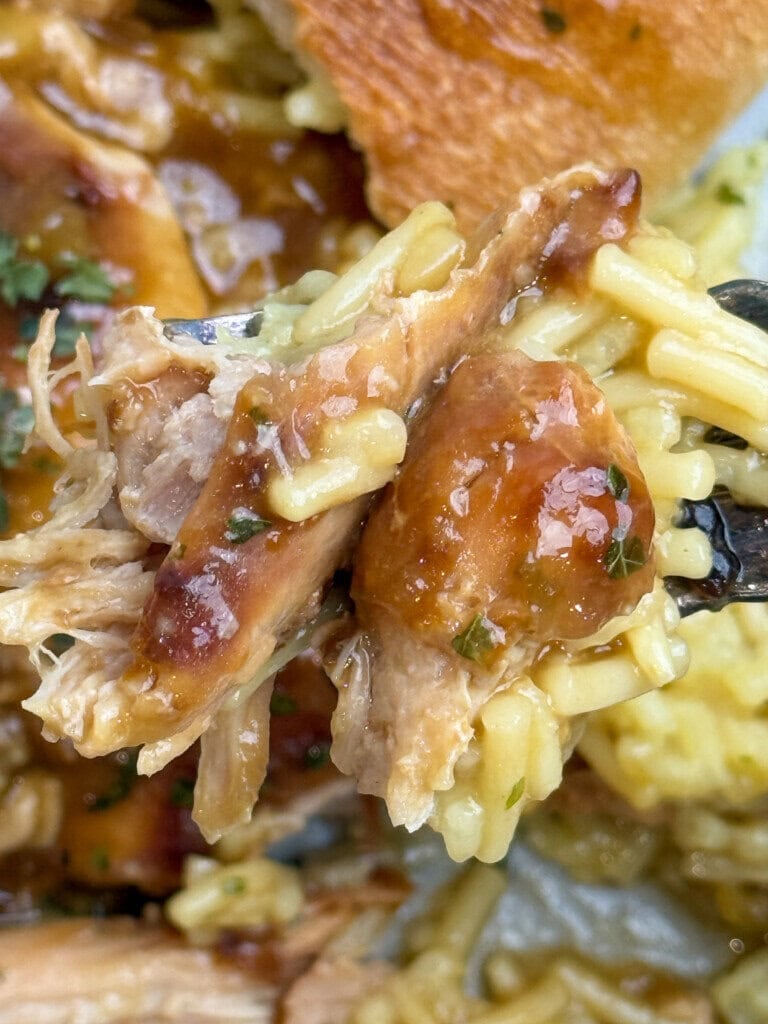 A close-up of a forkful of shredded chicken with creamy yellow rice, topped with brown gravy, with more rice and a piece of bread visible in the background.