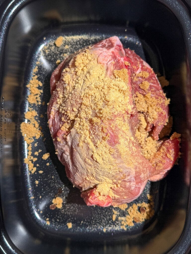 A piece of raw pork in a black roasting pan, sprinkled with a layer of light brown sugar and garlic powder.