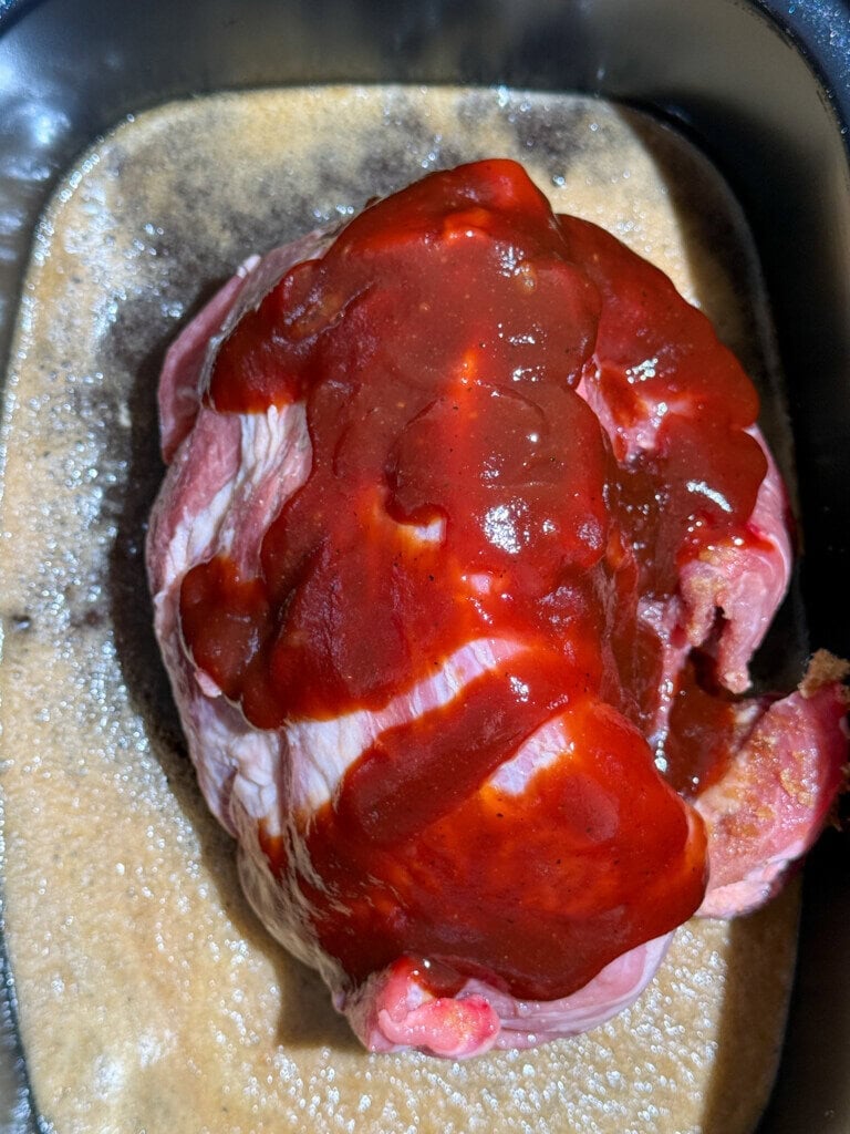 A raw piece of pork in a slow cooker, covered with reddish-brown barbecue sauce, sits in a foamy liquid Coca-Cola