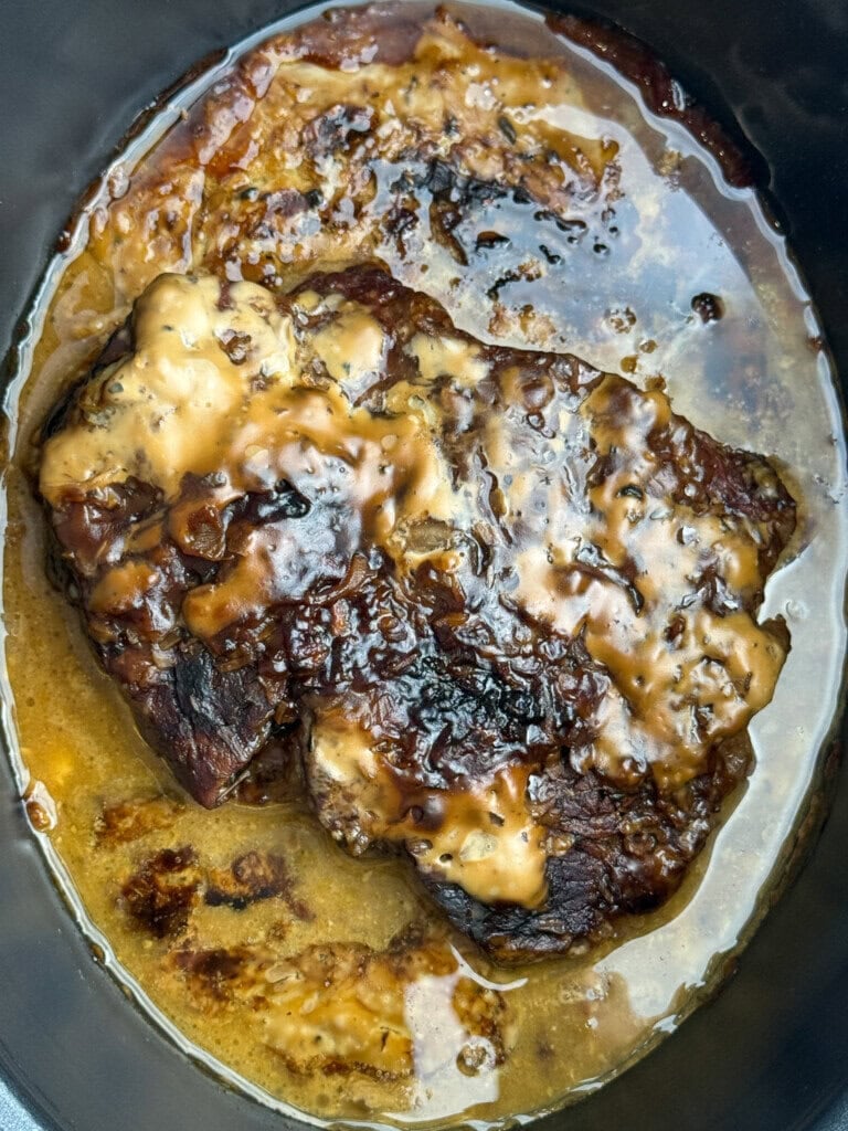 A cooked piece of meat with a browned, slightly charred surface and gravy sauce on top, sitting in a pool of juices inside a dark slow cooker or pot.