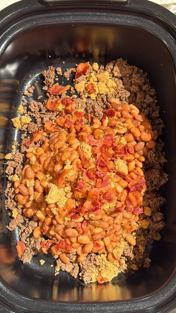 Ground beef, baked beans, crumbled bacon, and brown sugar layered in a black slow cooker, ready to be mixed and cooked. Sunlight highlights the ingredients.