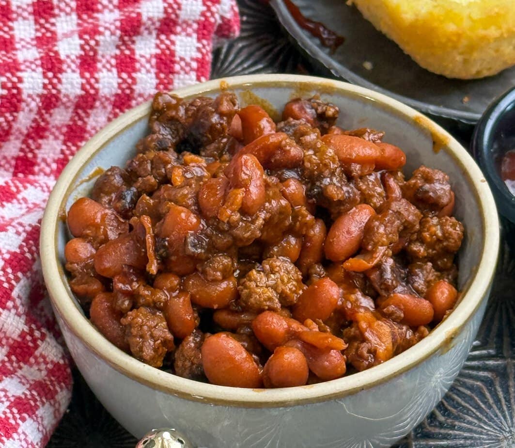 A bowl filled with baked beans and ground meat sits on a table next to a red-and-white checkered towel, with a plate of cornbread partially visible in the background.