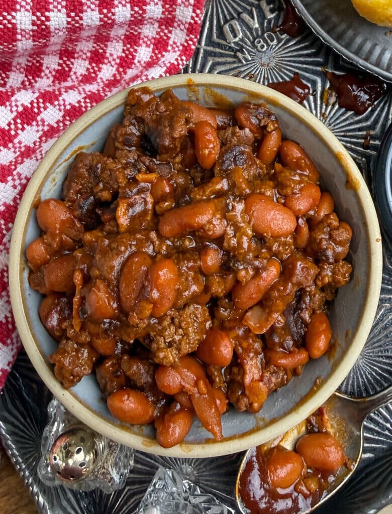 A bowl of baked beans mixed with ground beef in a rich, reddish-brown sauce, placed on a metal tray next to a red and white checkered cloth. A spoon with a bit of the mixture is nearby.