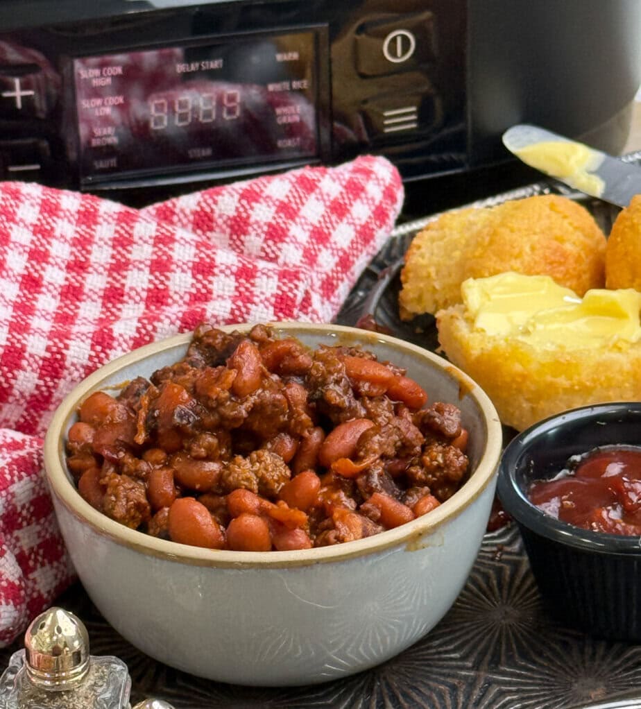 A bowl of chili with beans and ground meat sits in front of a slow cooker. Nearby are buttered cornbread muffins, a small dish of red sauce, and a red-and-white checkered cloth.