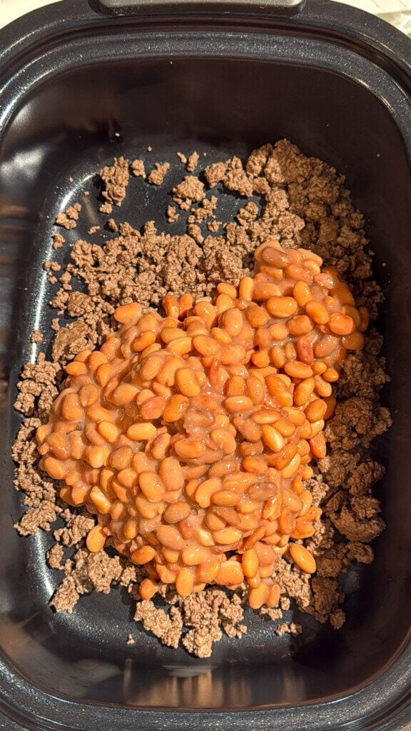 A black slow cooker contains a layer of cooked ground beef topped with baked beans in tomato sauce, ready to be mixed or cooked further.