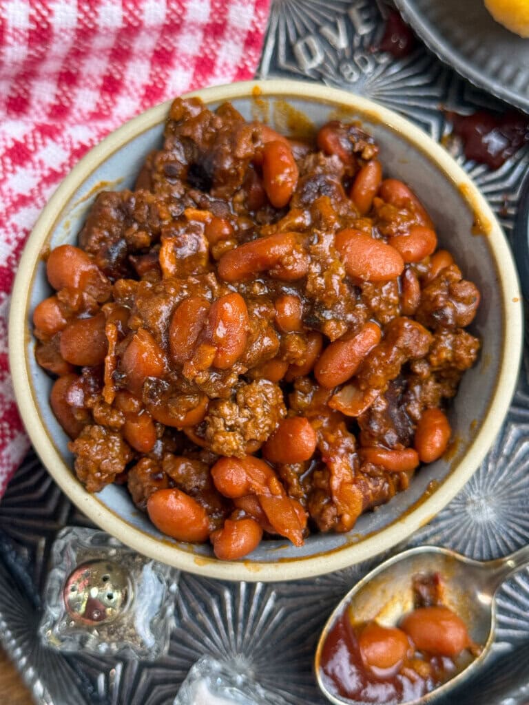 A bowl of chili with pinto beans and ground beef, placed on a metal tray next to a spoon, salt shaker, and a red-and-white checkered cloth.