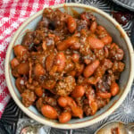 A bowl filled with baked beans and ground beef in a rich sauce sits on a metal tray next to a red and white checkered napkin, a salt shaker, and a small dish with some sauce.