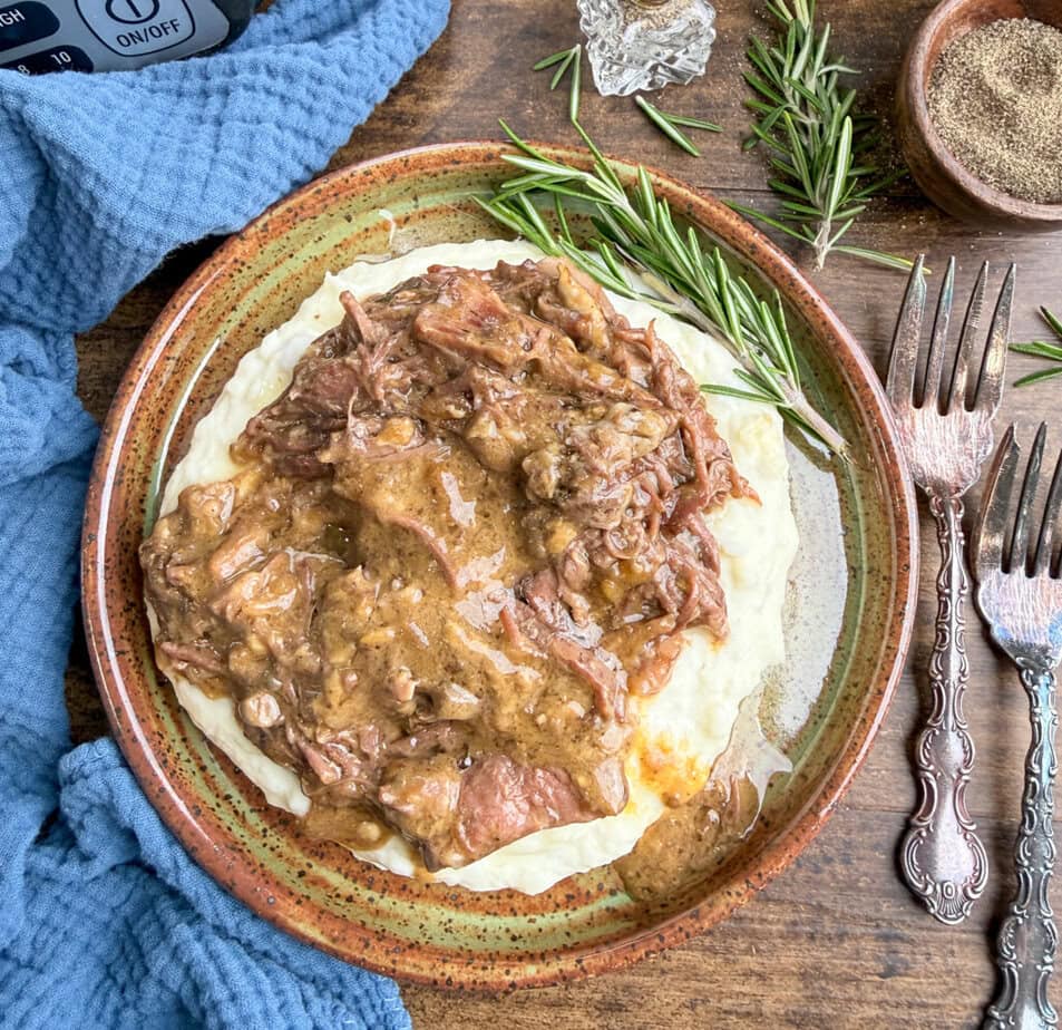A plate of shredded beef with brown gravy served over mashed potatoes, garnished with a sprig of rosemary, sits on a rustic wooden table beside two vintage forks, a blue cloth, and a bowl of ground pepper.