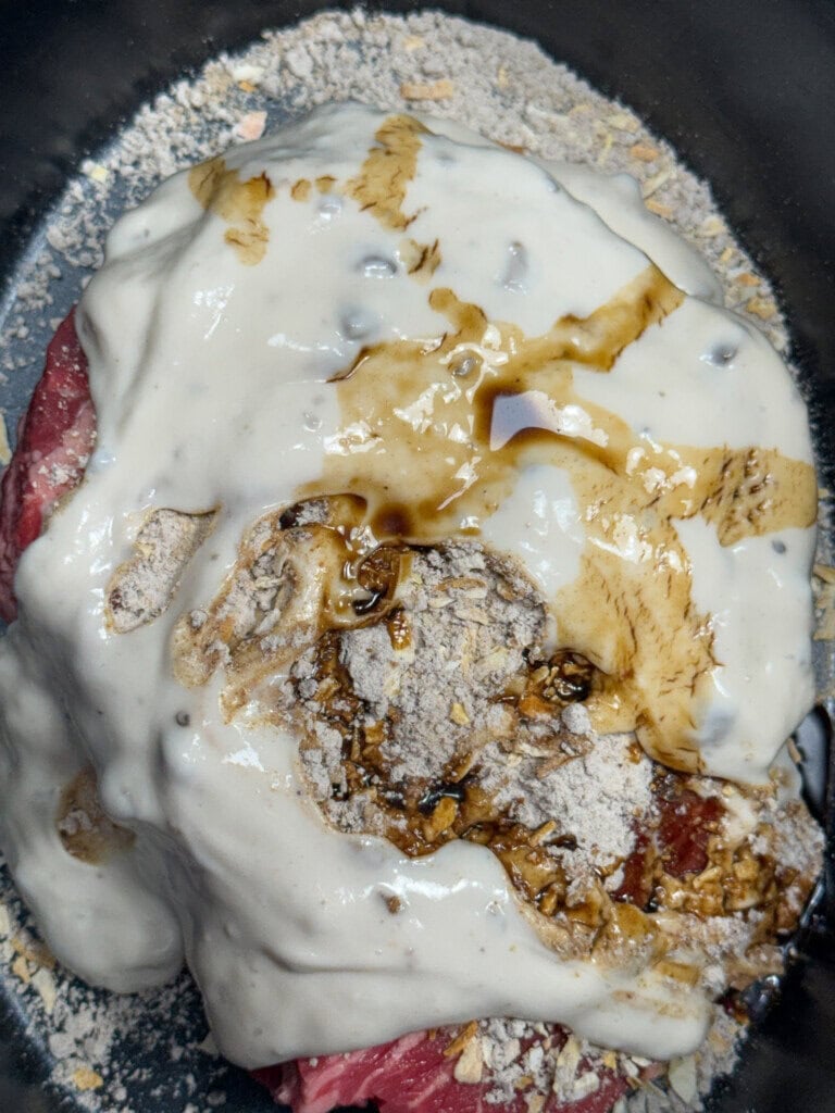 Raw beef covered with a creamy mushroom soup, onion soup mix and gravy mix, and a drizzle of Worcestershire sauce in a pot.