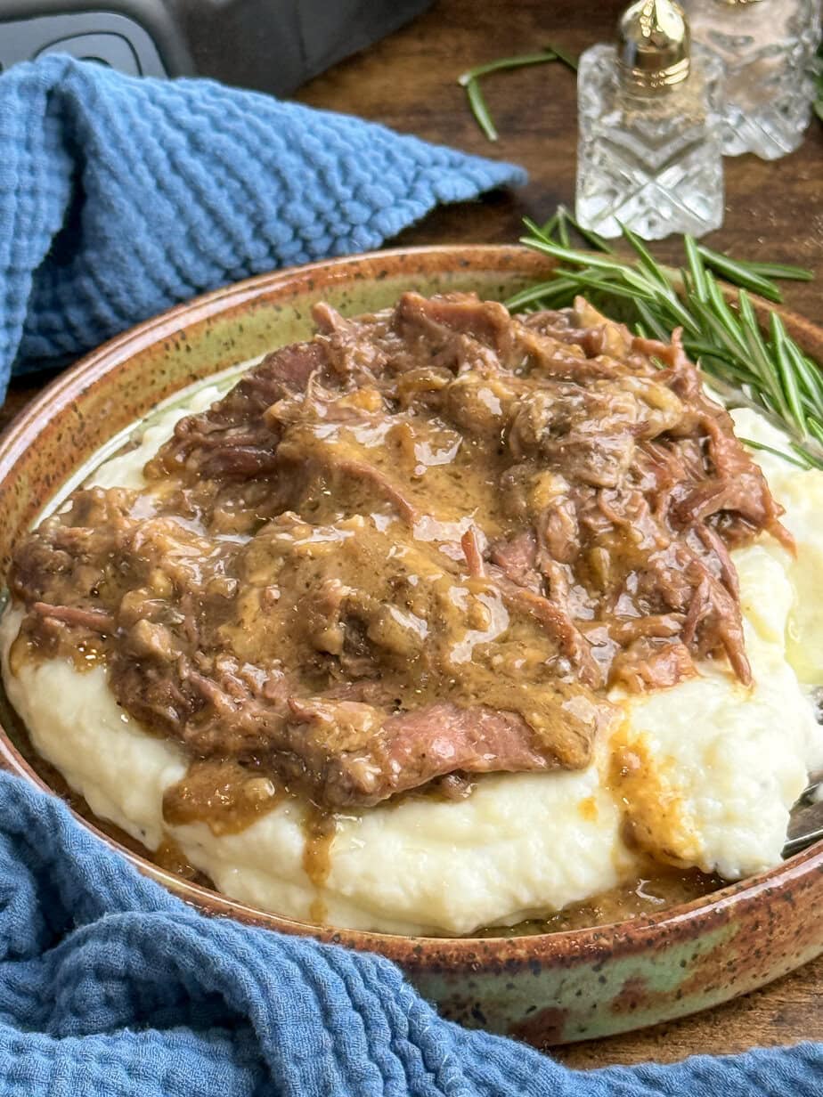 A plate of creamy mashed potatoes topped with shredded beef and rich brown gravy, garnished with a sprig of fresh rosemary, sits on a wooden table with a blue textured cloth nearby.