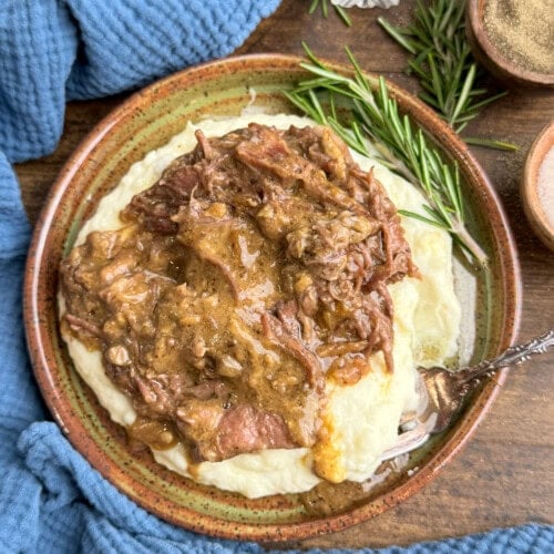 A bowl of creamy mashed potatoes topped with tender shredded beef and rich brown gravy, garnished with fresh rosemary, sits on a wooden table next to a blue cloth and small bowls of seasoning.