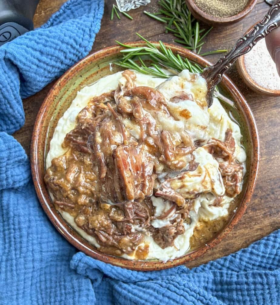 A bowl of creamy mashed potatoes topped with shredded beef and rich brown gravy, garnished with fresh rosemary, sits on a wooden surface next to a textured blue cloth.