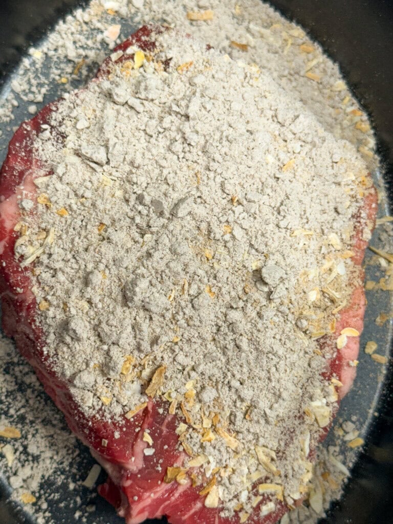 A raw piece of meat in a pan is covered with a layer of dry seasoning mix that appears to include powdered ingredients and dried onion flakes.