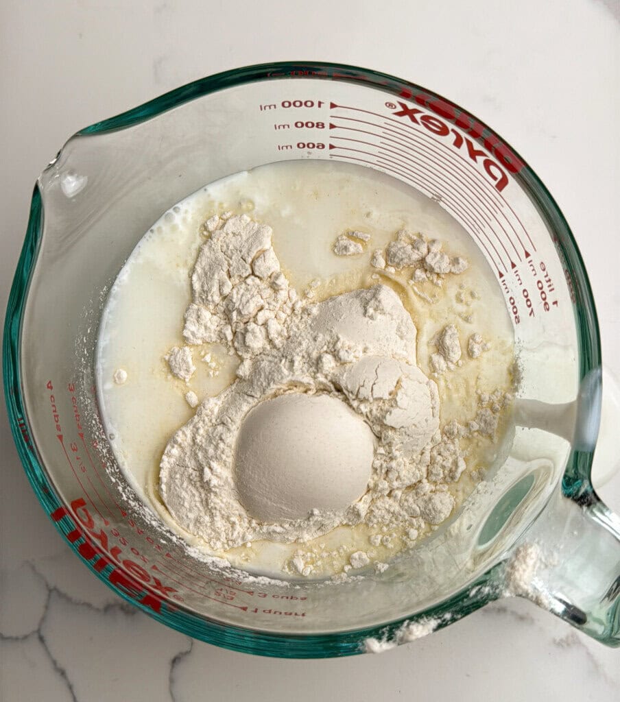 A glass measuring cup filled with flour, buttermilk, and water sits on a white countertop, ready to be mixed for baking.