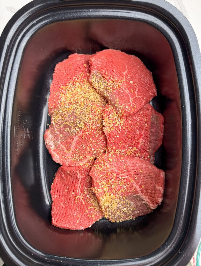 Six raw cube steaks are placed in a black slow cooker, sprinkled with mixed onion. The meat appears fresh and ready for cooking.
