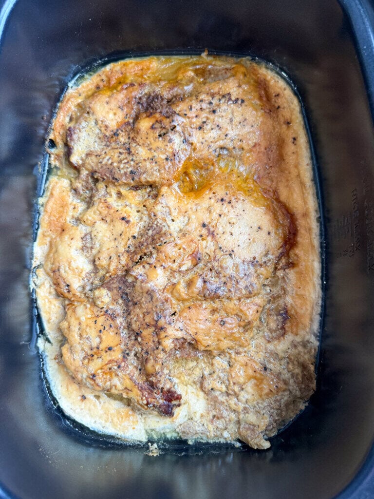 A close-up of a cooked cube steak in a black slow cooker, topped with a browned, seasoned crust and visible juices around the edges.