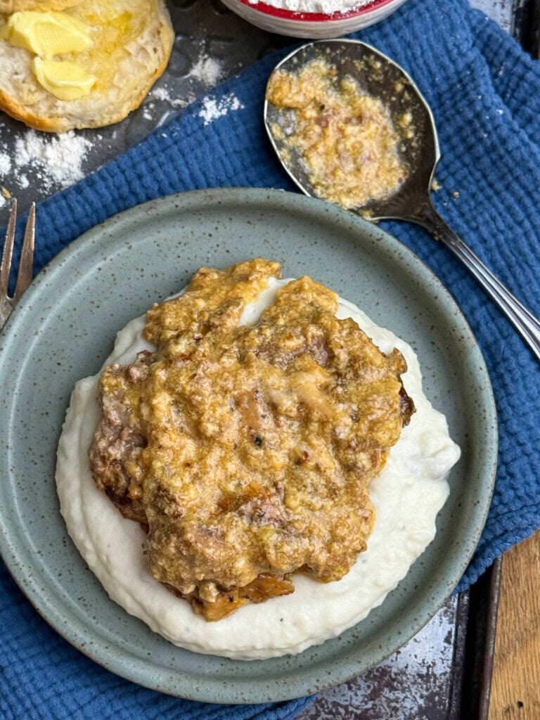 A plate with a serving of white mashed potatoes topped with a browned, creamy meat gravy. A spoon with more gravy and a buttered biscuit sit nearby on a blue cloth.