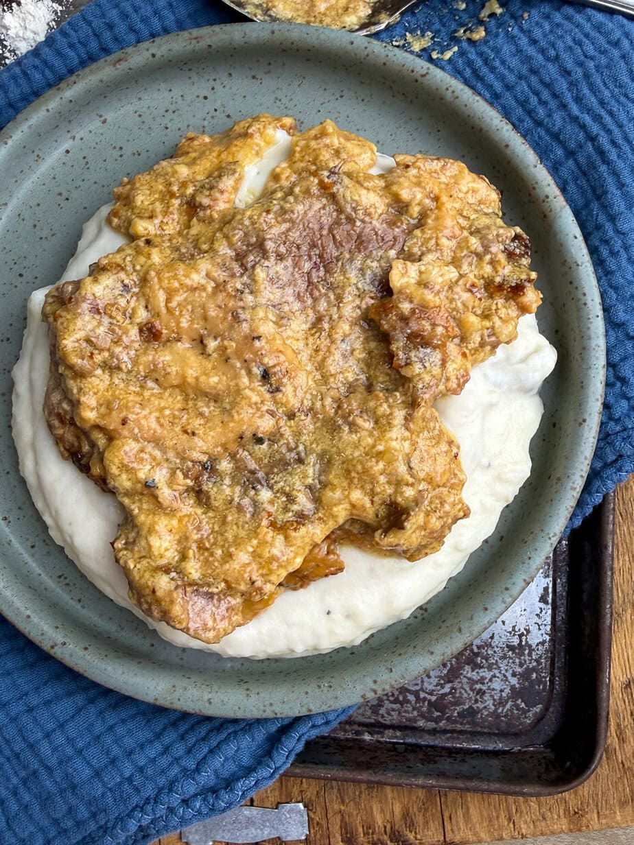 A plate with a serving of creamy mashed potatoes topped with a golden-brown piece of cube steak, all set on a blue textured cloth.