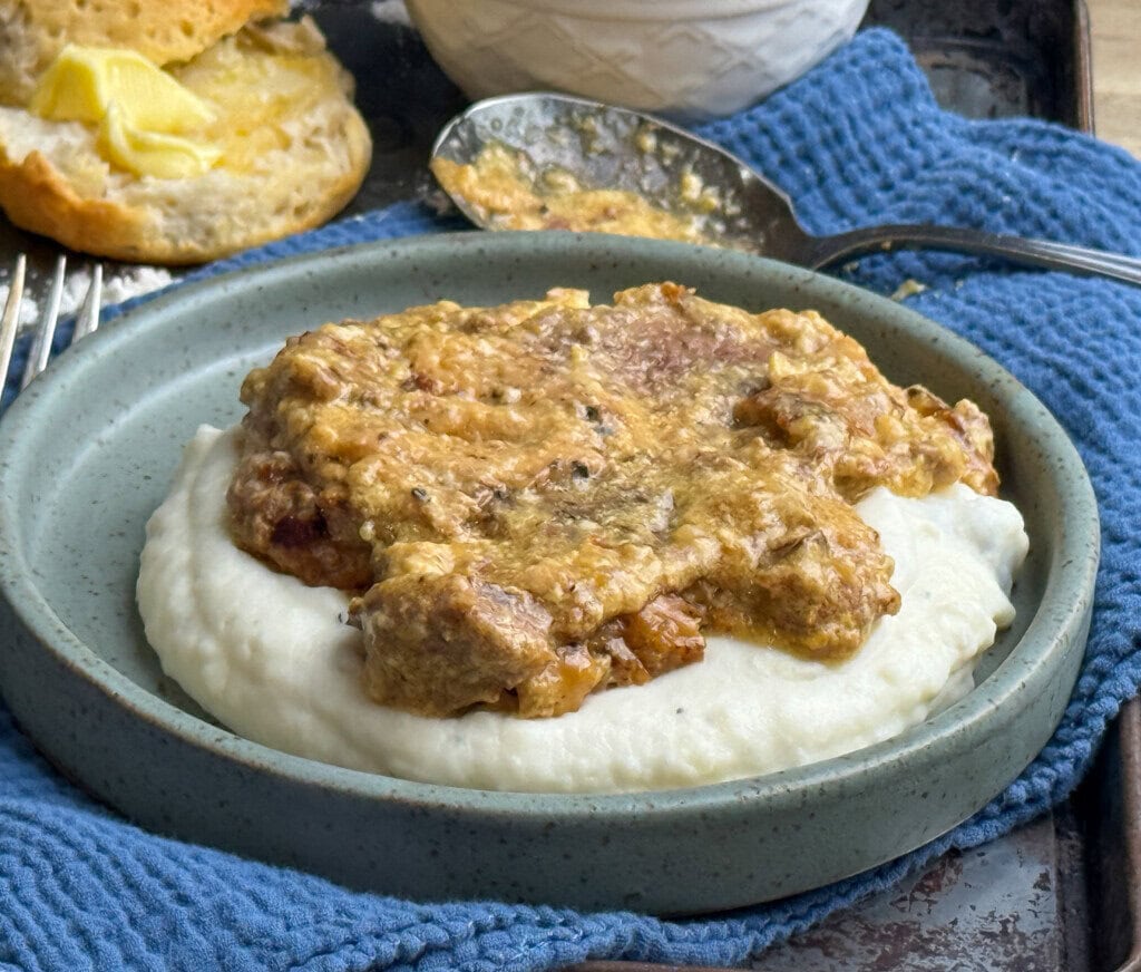 A plate of creamy mashed potatoes topped with thick, brown cube steak and buttermilk gravy sits on a blue cloth, with a spoon and a buttered biscuit in the background.