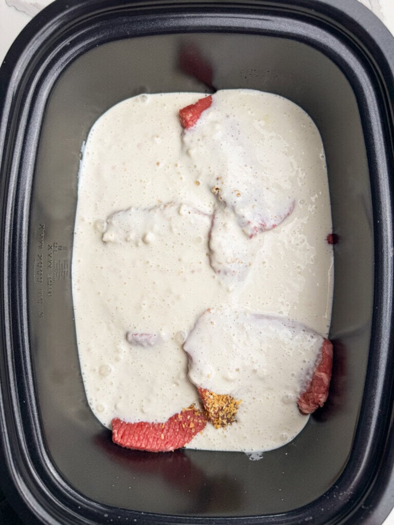 Raw beef steaks in a black slow cooker insert, partially covered with a thick, creamy white buttermilk gravy and sprinkled with seasonings.