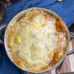 A close-up of a bowl of French onion soup, topped with melted, golden-brown cheese, sitting on a blue cloth next to a slice of bread and a spoon on a wooden surface.