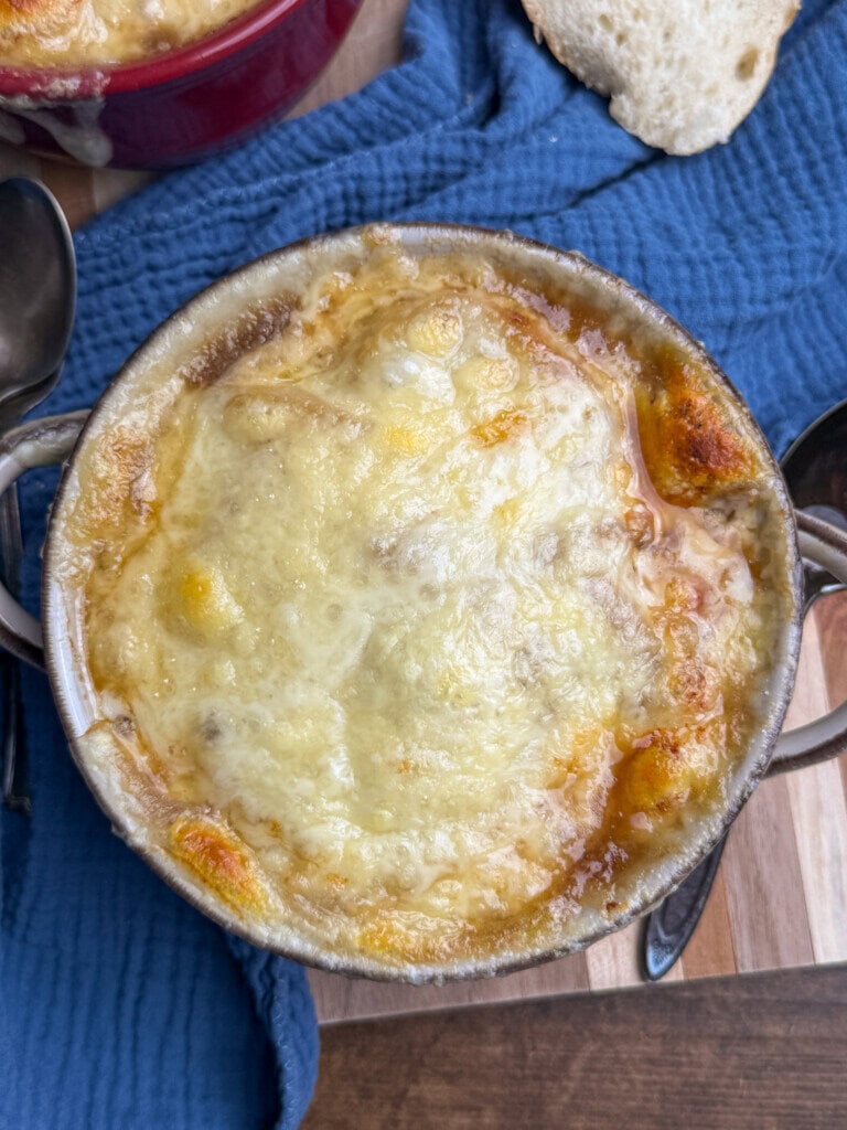 A close-up of a bowl of French onion soup, topped with melted, golden-brown cheese, sitting on a blue cloth next to a slice of bread and a spoon on a wooden surface.