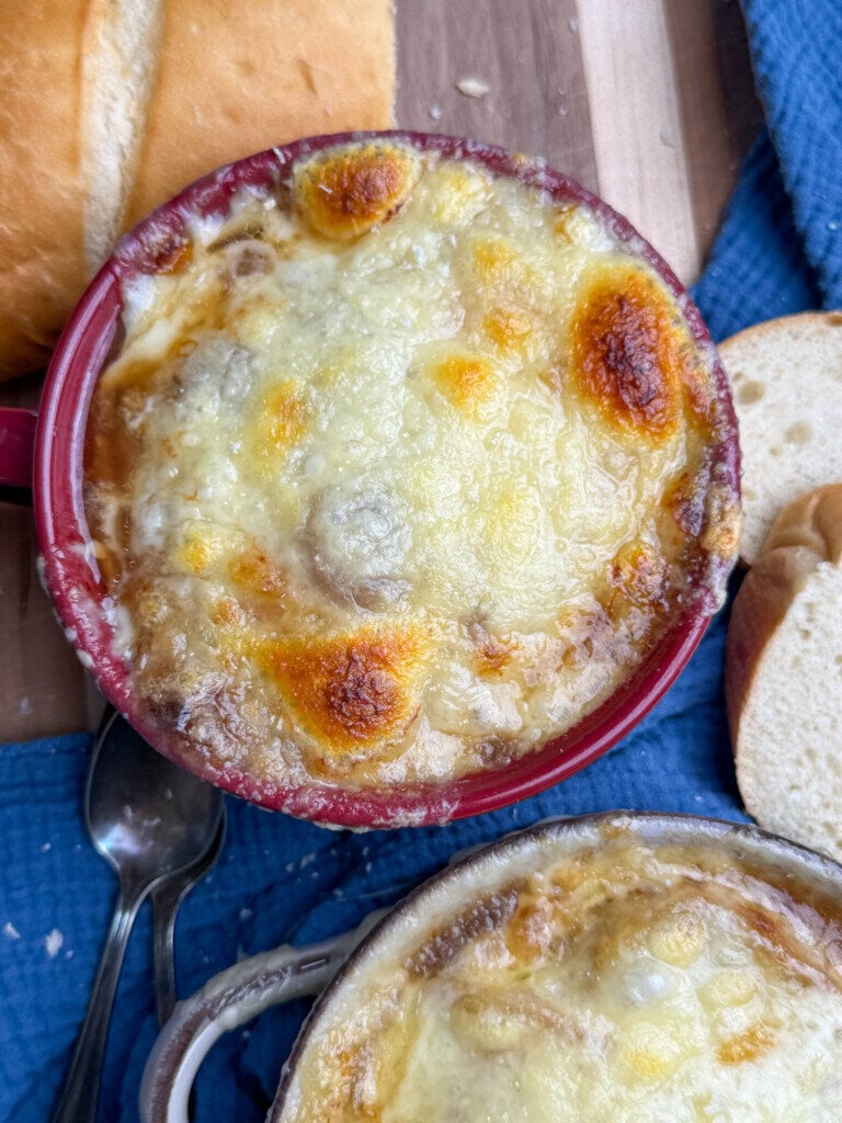 A bowl of French onion soup topped with melted, golden-brown cheese sits on a wooden surface next to slices of bread, a blue cloth, and metal spoons.