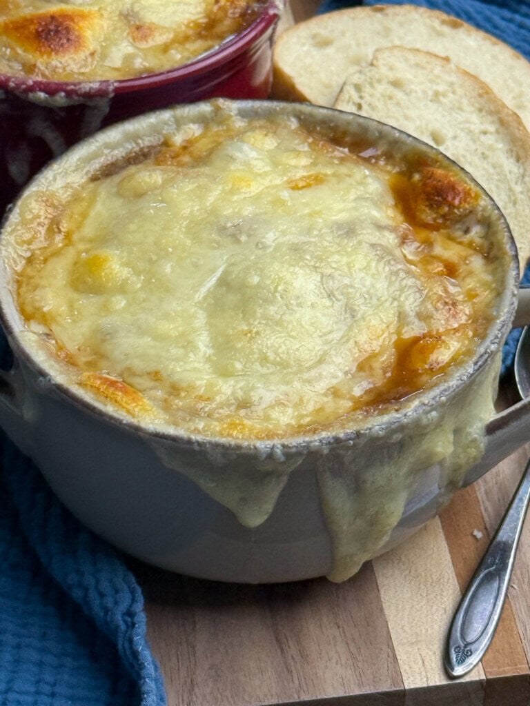 A bowl of French onion soup topped with melted, golden-brown cheese, with cheese dripping over the sides. Slices of toasted bread and a spoon are next to the bowl on a wooden surface.