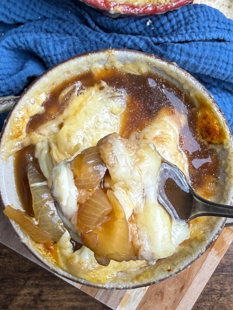 A close-up of French onion soup in a bowl, topped with melted cheese and toasted bread, with a spoon lifting a cheesy, onion-filled portion over the bowl. A blue cloth is in the background.