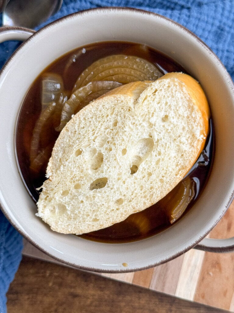 A bowl of French onion soup topped with a slice of crusty bread, with sliced onions visible in the dark broth, placed on a blue cloth and wooden surface.