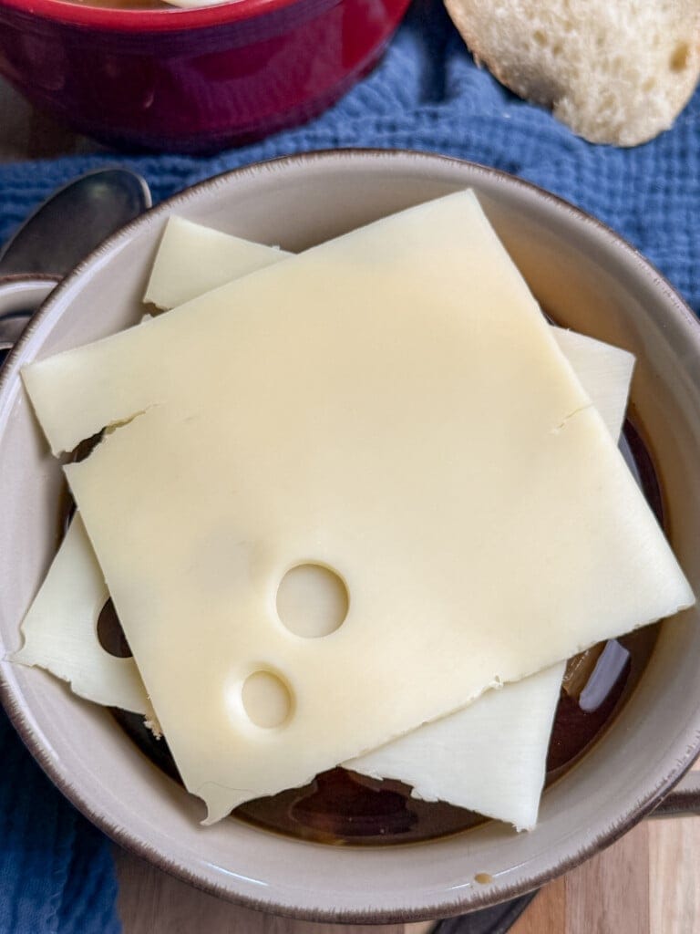 A bowl containing dark broth topped with several slices of Swiss cheese, with a piece of bread and a blue cloth in the background.