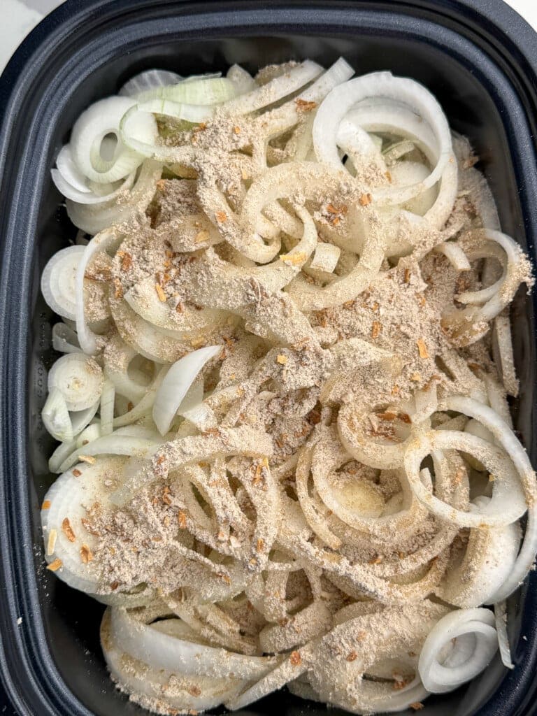Sliced white onions covered with a layer of dry seasoning and breadcrumbs in a black rectangular dish, ready for cooking.