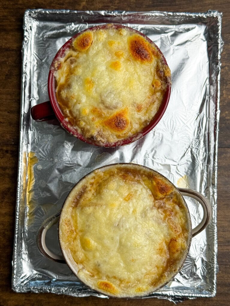 Two bowls of French onion soup topped with melted, slightly browned cheese sit on a foil-lined baking sheet. The bowls are placed side by side on a wooden table.
