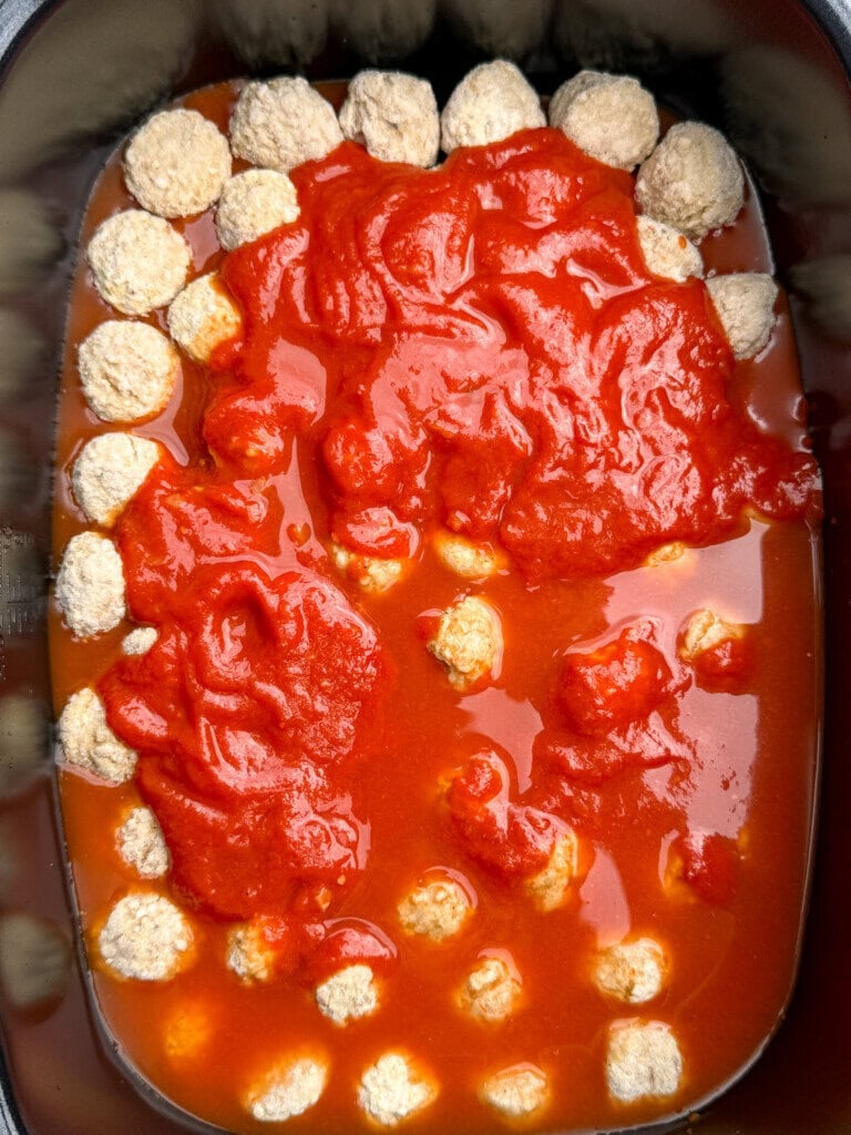 Meatballs arranged around the edges and bottom of a black slow cooker, covered with chunky red tomato sauce.