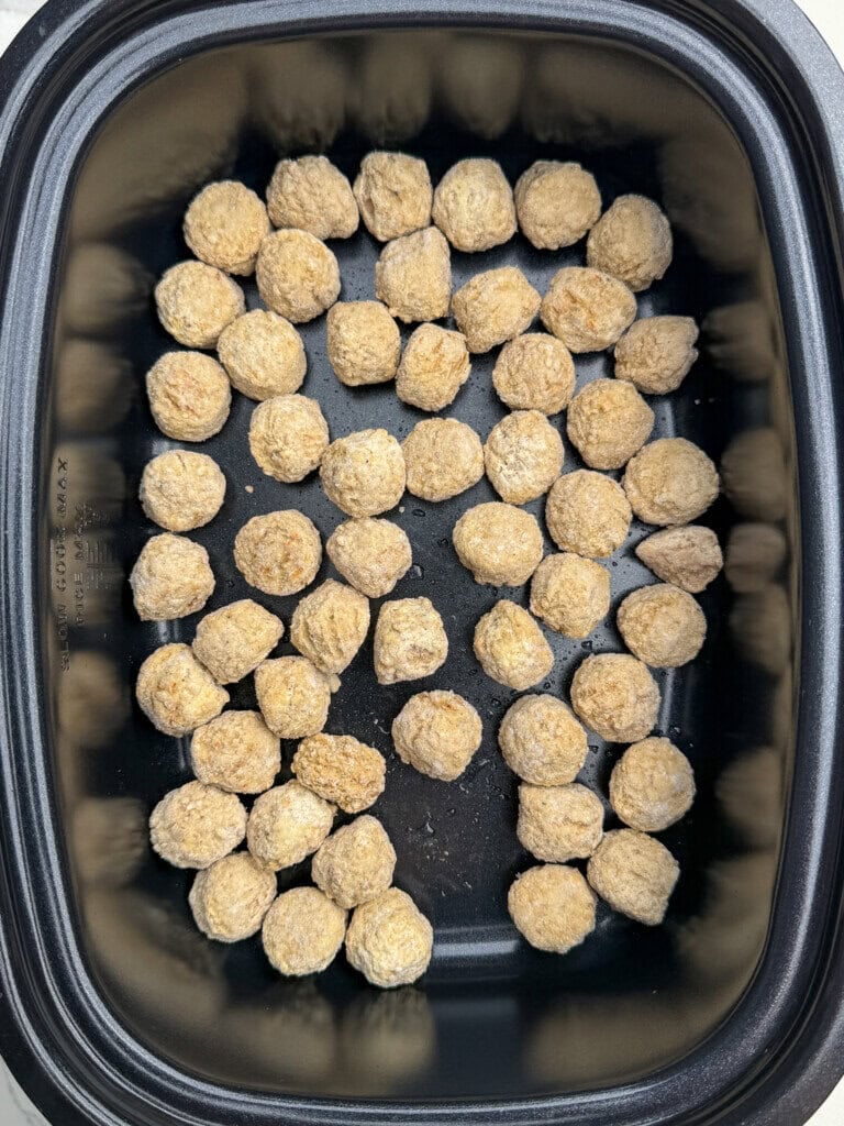 A black rectangular crock pot filled with evenly arranged, uncooked frozen breaded meatballs in neat rows.