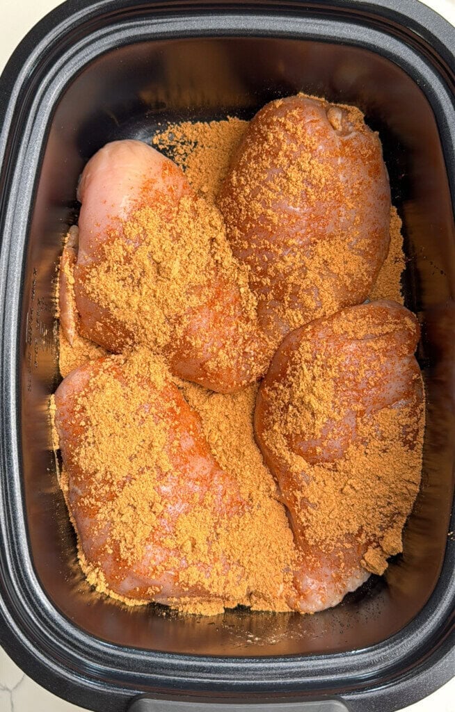 Four raw chicken breasts in a black slow cooker, covered with a generous layer of orange-brown seasoning or spice mix.