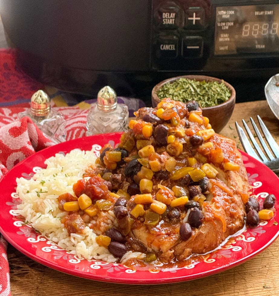A plate of rice served with chicken topped with a colorful mix of black beans, corn, and tomatoes, sitting in front of a slow cooker and a bowl of herbs on a wooden table.