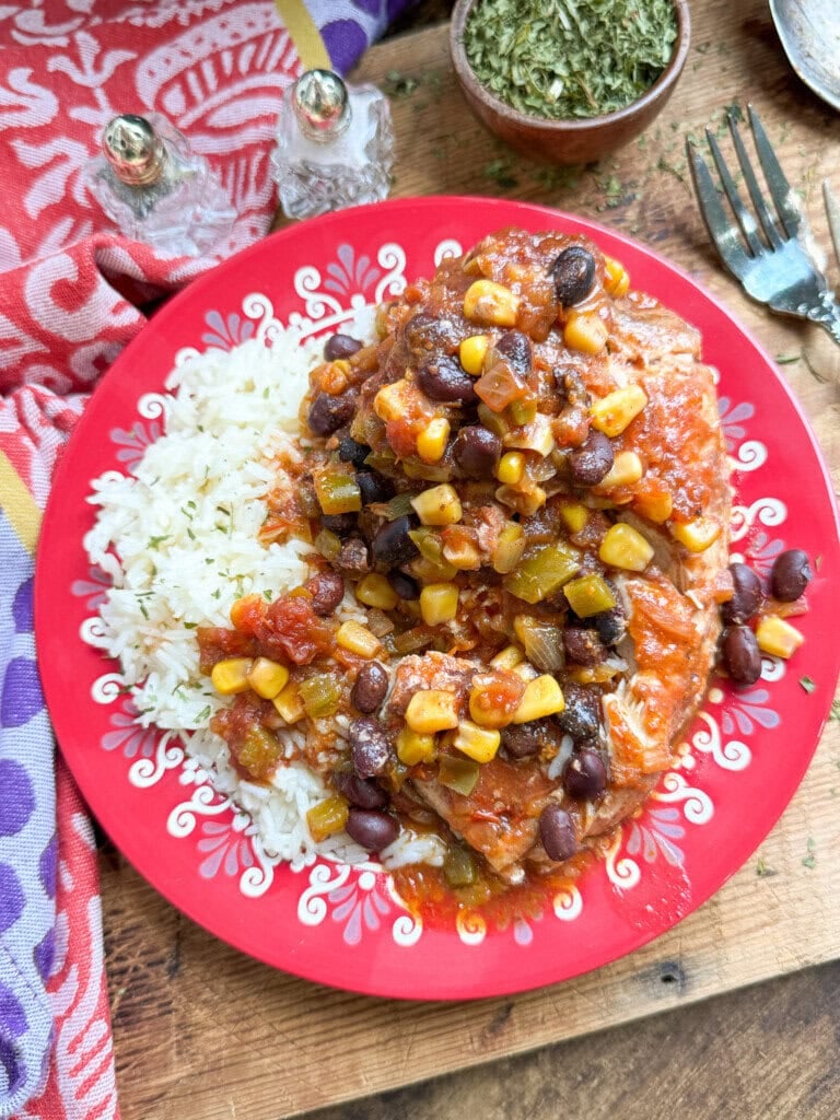 A red plate with white rice and a colorful chicken meal featuring corn, black beans, diced tomatoes, green chilies, and sauce, set on a wooden table with herbs, salt and pepper shakers, and a fork nearby.