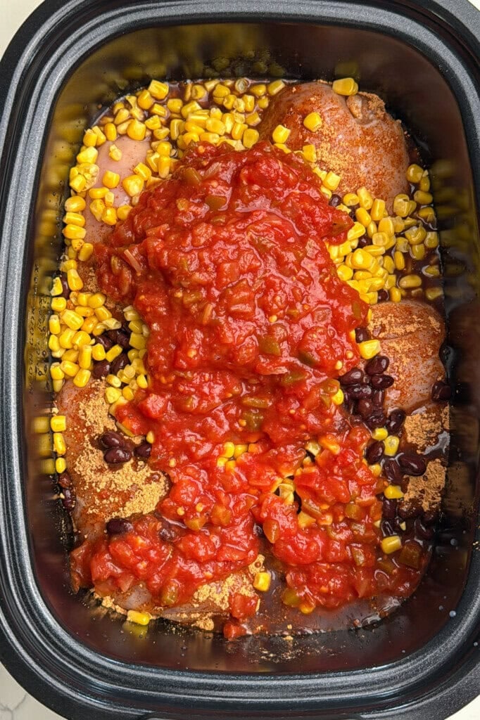 Chicken breasts in a slow cooker topped with black beans, corn, and chunky salsa, ready to be cooked.