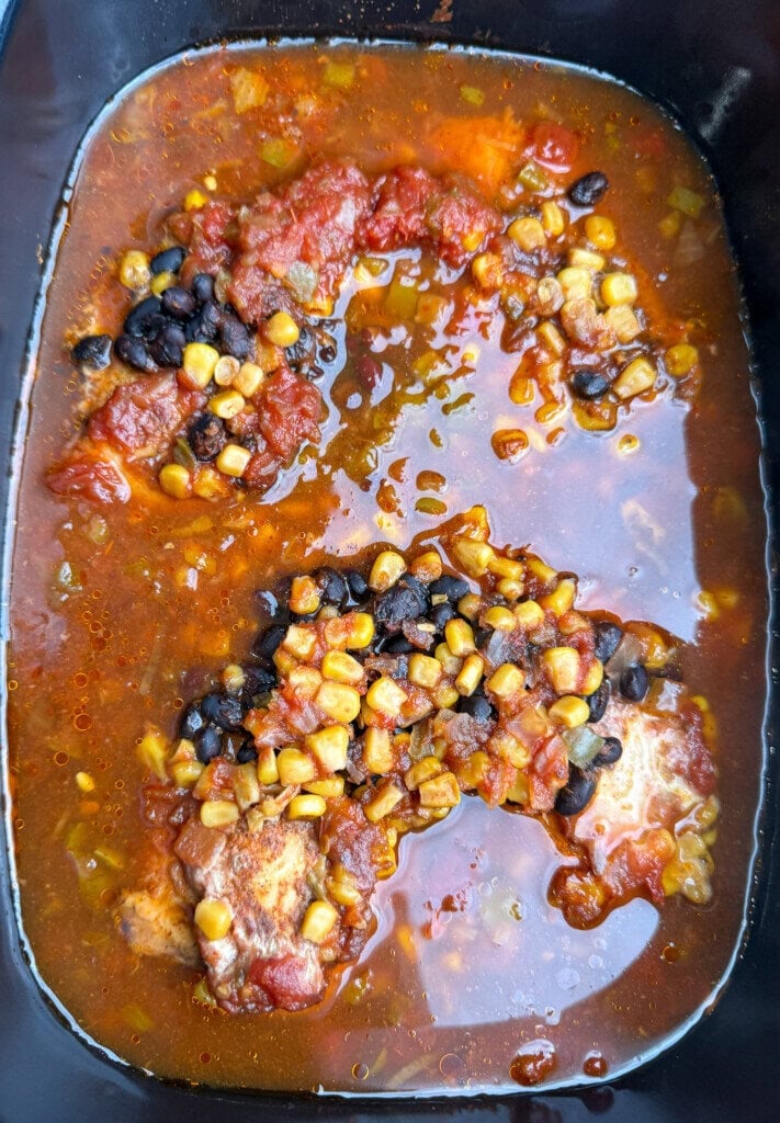 Chicken pieces in a slow cooker with a tomato-based sauce, topped with black beans and corn. The mixture appears to be simmering, with visible oil and spices in the liquid.