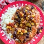 A colorful plate with white rice and a saucy mix of chicken, black beans, corn, tomatoes, and green peppers, served on a red floral-patterned dish. A fork and bowl of herbs are nearby on a wooden table.