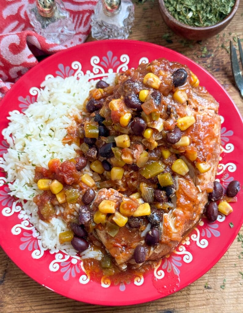 A colorful plate with white rice and a saucy mix of chicken, black beans, corn, tomatoes, and green peppers, served on a red floral-patterned dish. A fork and bowl of herbs are nearby on a wooden table.