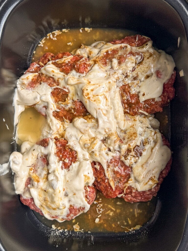 Ground beef topped with a creamy white mushroom soup, seasonings, and diced onions in a slow cooker, ready to be cooked.